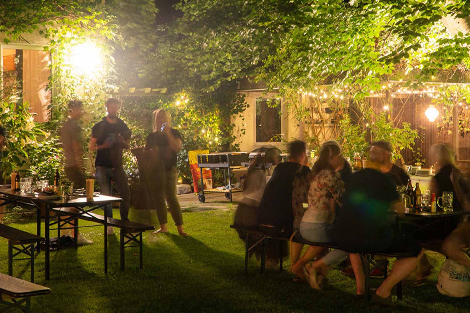 PINC Garten im dunkeln während einer Party. Menschen sitzen auf Bierbänken und unterhalten sich oder stehen mit einem Glas in der Hand da.