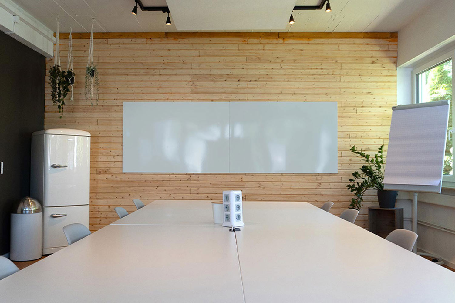 Langer Meetingtisch mit Stühlen und einem Whiteboard an einer Holzwand. Daneben sieht man einen Kühlschrank, Pflanzen und ein mobiles Flipchart. Im Anschnitt sieht man ein offenes Regal.